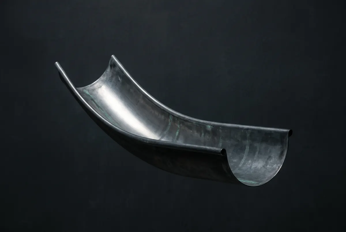 A length of half-round gutter in dark weathered zinc, a long crescent of light running the inside of its curve against a dark void.