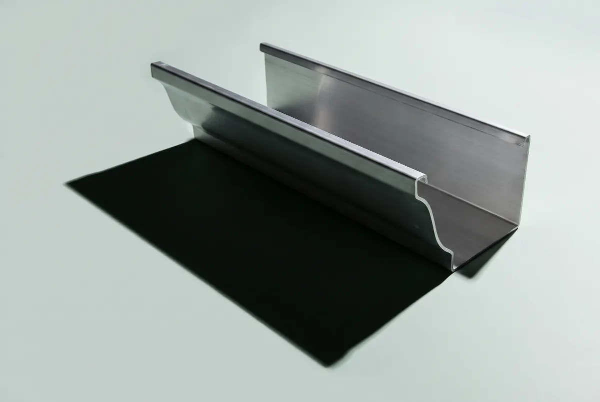 A short offcut of K-style gutter in mill aluminum on a pale surface, its ogee front lip throwing a hard shadow that repeats the curve.
